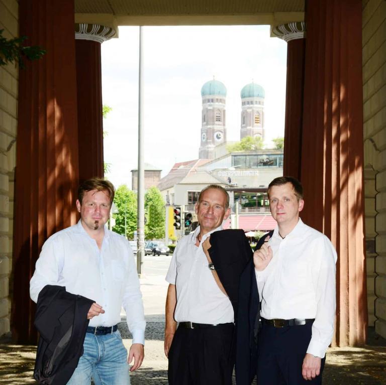 Drei Männer im Freien vor einer Münchner Stadtansicht mit Bäumen und Türmen